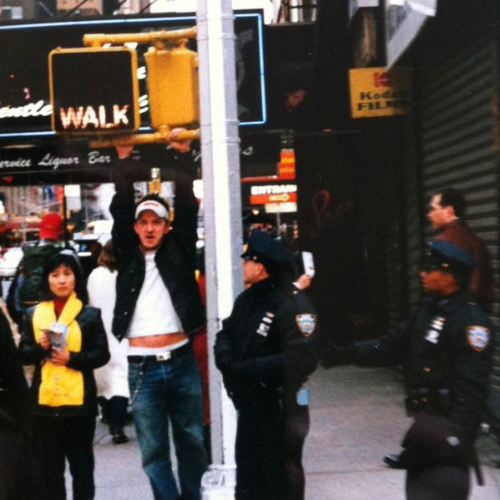An image of a person hanging from a street lamp in a city that looks like New York. The photo has a distinctly 90s feel.