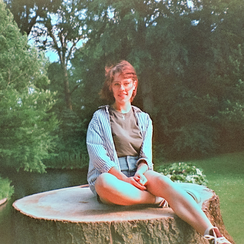 A photograph of a person sat on a tree stump. They wear double denim and their hair flows in the wind. The photo has a retro feel.
