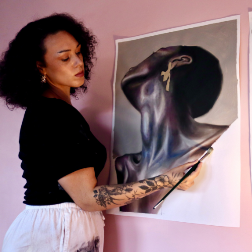 An artist stands in front of a piece of artwork against a pink wall. They apply brush strokes to the artwork and appear deep in concentration.