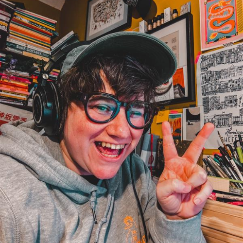 A person with dark, short hair wearing glasses, a baseball cap and headphones smiles at the camera and pulls a peace sign.
