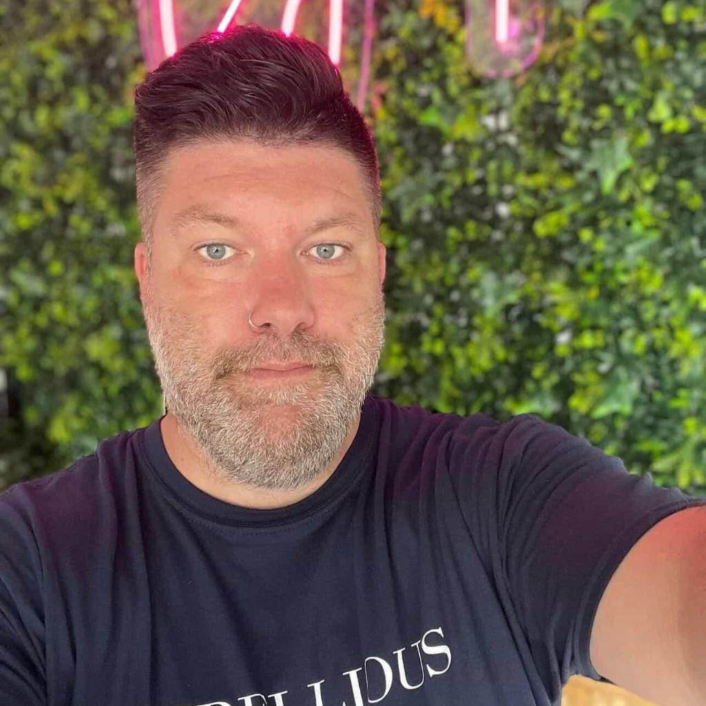 A selfie of a person with short, dark hair and a beard. Wearing a black t-shirt and stood against a green wall.