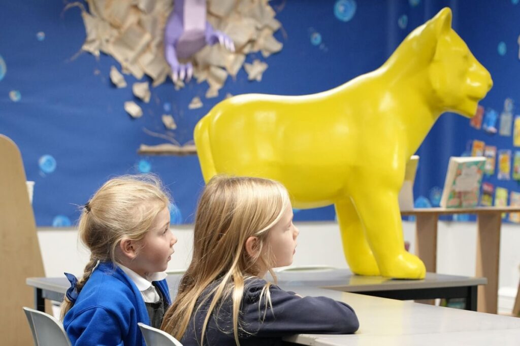 An image of two school children listening intently to a teacher while a bright yellow lion cub stands proud in the background.