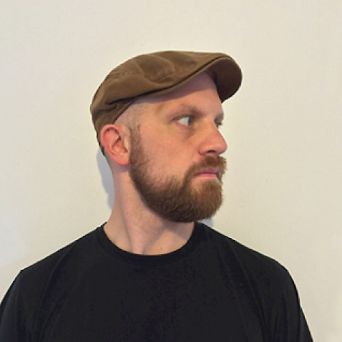 A person wearing a brown flatcap and a black tshirt stands against a plain wall and looks off to the left.