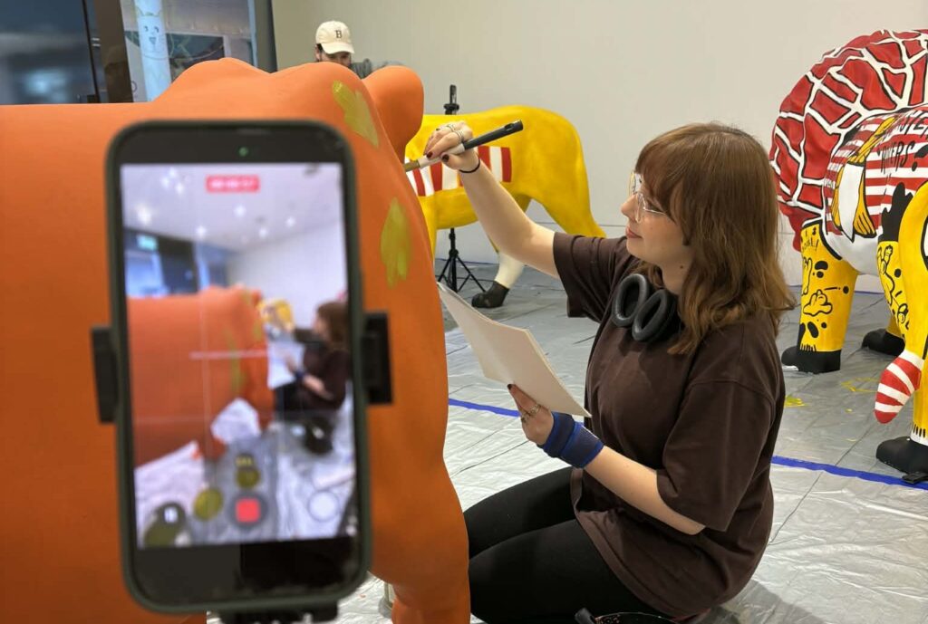 Artist Christy Johnson works on her bright-orange lioness sculpture in focus in the background, in the foreground her phone records.