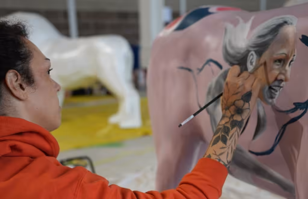 A photograph of a person painting a sculpture. They are painting a portrait of an athlete on the side of a pink sculpture.