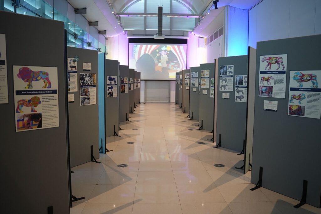 A photograph of an event for Pride of Yorkshire. It's in a gallery, and shows a long room with both sides showing designs for the trail. In the distance, a video of artists painting plays with ambient lighting either side.