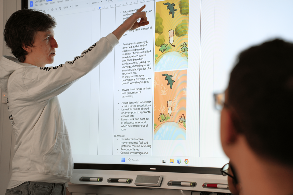 A photograph of a person in a white hoodie pointing at a whiteboard - on the board is a game design document. In the foreground, someone watches on.