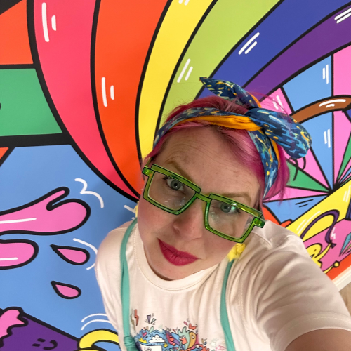 A photograph of a person with pink hair and bright green square glasses stood in front of a rainbow mural.