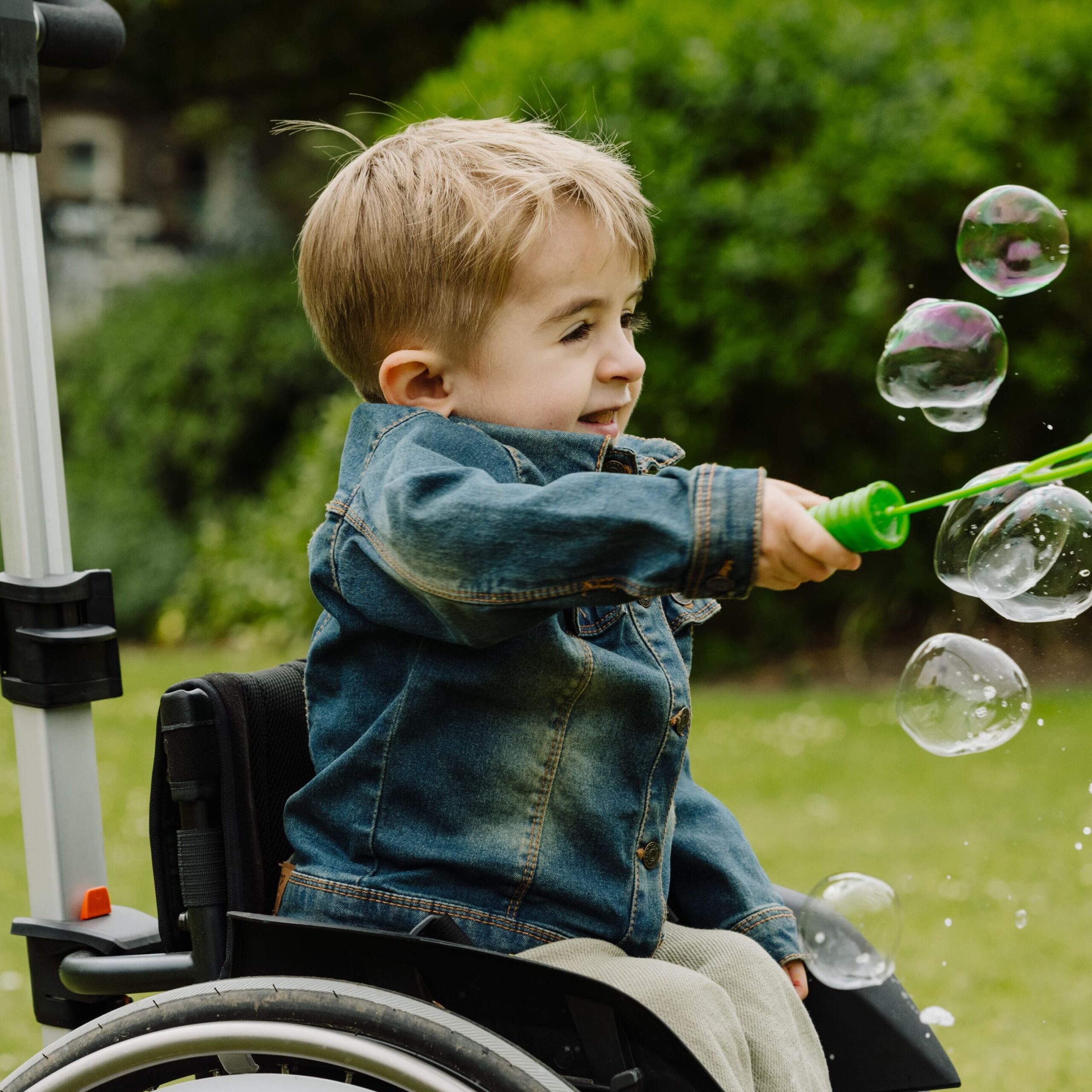 A child in a wheelchair holds a bubble wand and points it towards the right - bubbles are all around and the child is smiling.