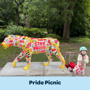 A small child smiles gleefully next to a brightly painted lioness sculpture that says STAY PROUD in big, pink letters. They are in a park and the background is made of lush, green trees. In front of them, a banner reads 'Pride Picnic'