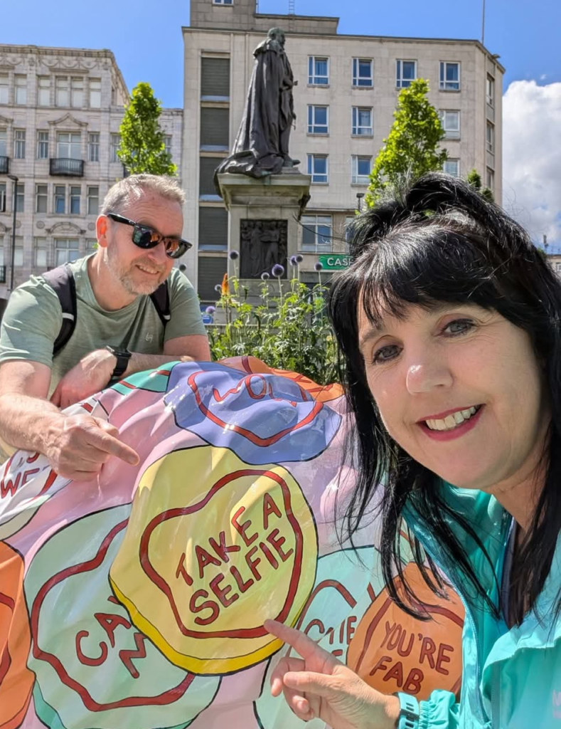 Two people stand on a sunny day with a bright pink lion sculpture covered in love heart sweets. They point at one that says 'take a selfie'.