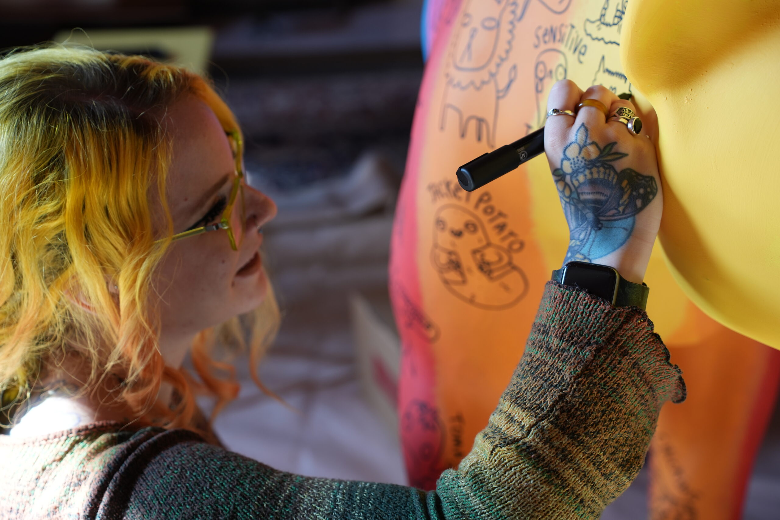 A person with bright yellow and orange hair uses a pen on a brightly coloured lioness sculpture.