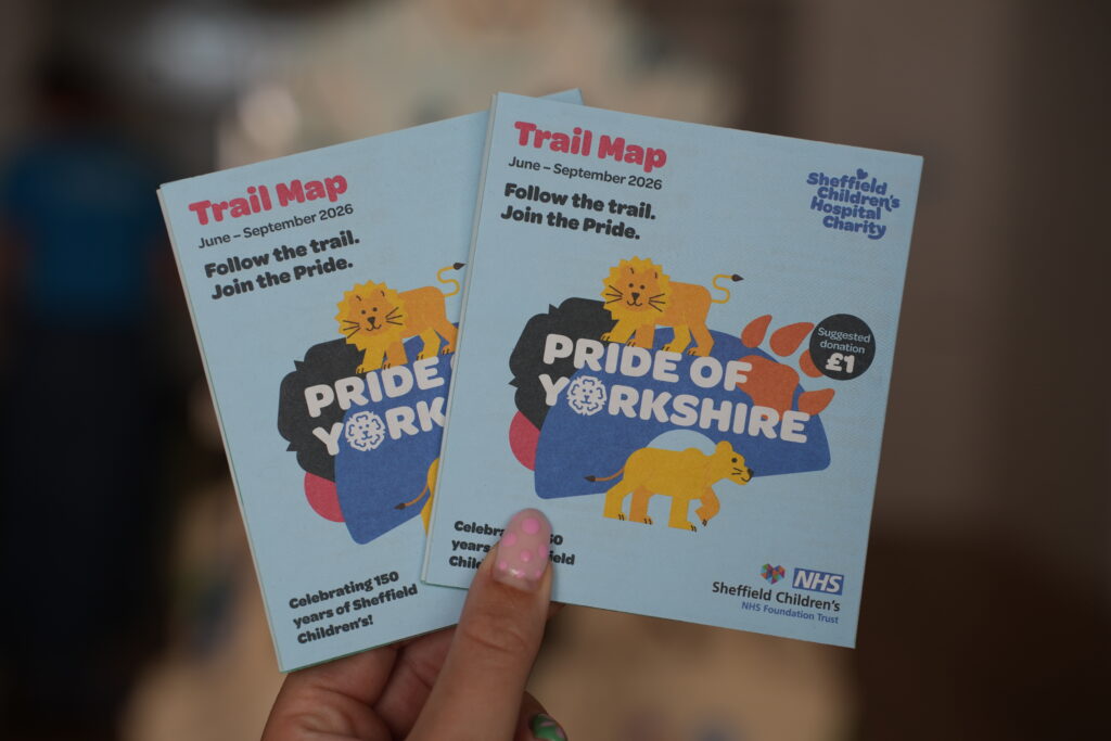 A hand holds two Pride of Yorkshire trail maps, these are small and square and say 'Pride of Yorkshire trail map' on the front.
