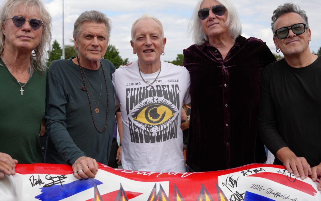 A photograph of rock band Def Leppard - which is five members - stood with their arms around each other in front of a Pride of Yorkshire lioness, with a union jack across the back.