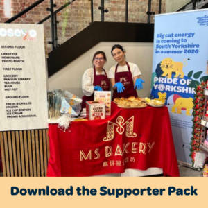 Two people wear aprons and blue gloves as they stand in a supermarket behind a table that reads 'MS Bakery' - they are fundraising for Pride of Yorkshire. In front of them, a banner reads 'Download the Supporter Pack'.