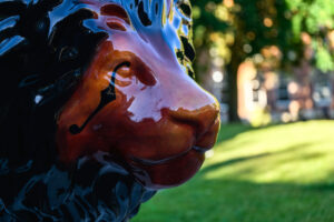 A lion sculpture painted in glossy dark brown with a black mane and small markings around the eyes. The photo is a close up of the face, with lush green trees in the background.