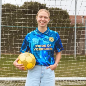 A blonde model wears a blue football jersey that reads PRIDE OF YORKSHIRE, with an embroidered lion face in the corner.